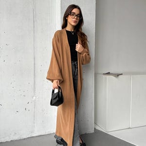 May include: A long, camel-coloured cardigan with a ribbed texture. The cardigan is worn over a black top and grey jeans. A small, black handbag is held in the hand. The person is wearing black shoes.