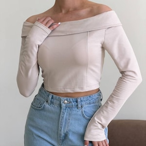 Può includere: Un top corto beige a maniche lunghe con scollo a barchetta, dettaglio del colletto ripiegato e cuciture verticali sul davanti, abbinato a jeans in denim azzurro a vita alta con un bottone in metallo argentato.