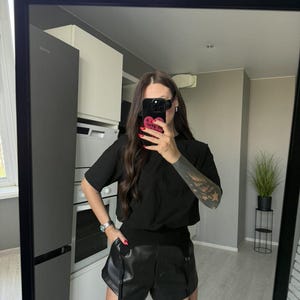 May include: Black t-shirt with a pocket and black leather shorts with suspender straps. The shorts are paired with black thigh-high stockings. The outfit is reflected in a large black framed mirror.