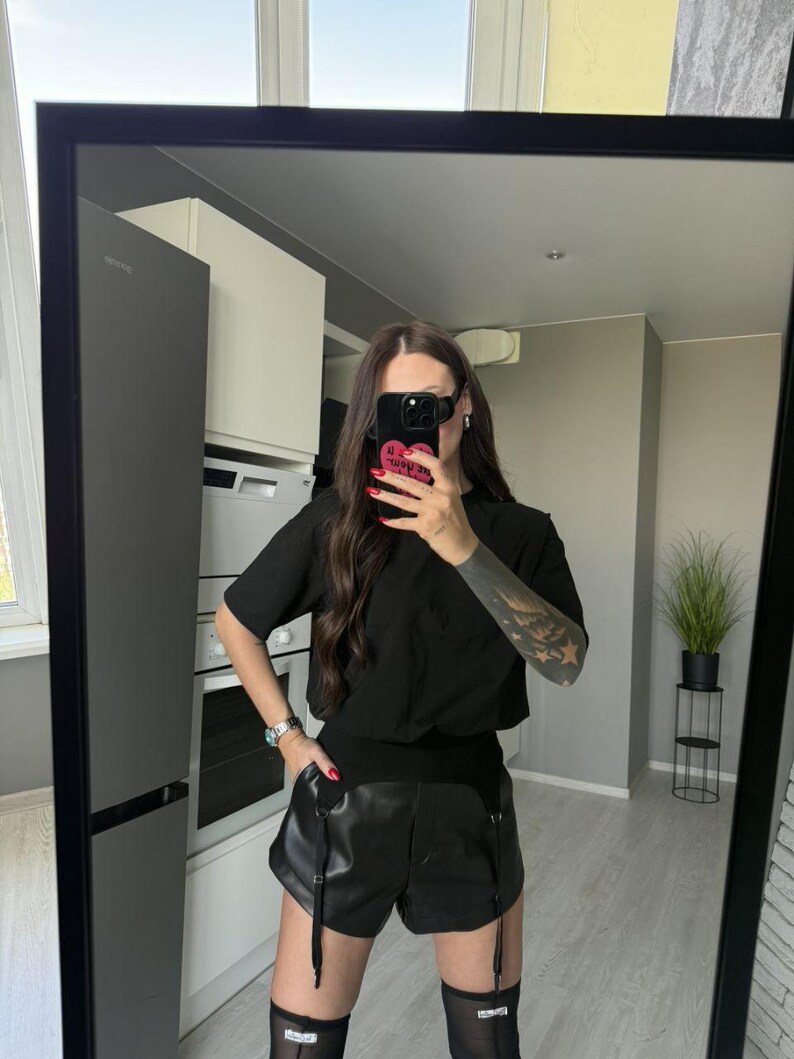 May include: Black t-shirt with a pocket and black leather shorts with suspender straps. The shorts are paired with black thigh-high stockings. The outfit is reflected in a large black framed mirror.