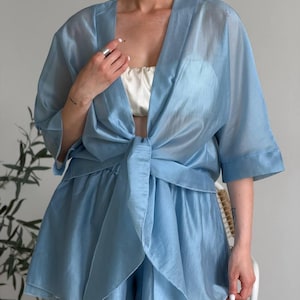 Op de afbeelding: Een lichtblauwe, doorschijnende tweedelige outfit. De set bestaat uit een top met korte mouwen in wikkelstijl, vastgebonden in de taille, en bijpassende shorts. Een witte top is eronder zichtbaar. De outfit is gemaakt van een lichte, vloeiende stof.