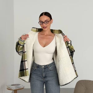 May include: A long-sleeved plaid shirt jacket featuring a green, black, and white checkered pattern with a thick white sherpa fleece lining. The garment is worn over a plain white v-neck bodysuit and paired with blue denim jeans and dark-rimmed glasses.