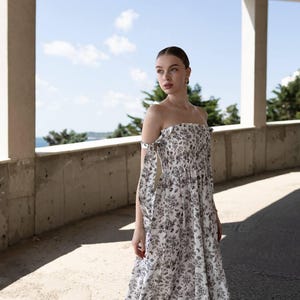 May include: A white, off-the-shoulder dress with a black floral print. The dress features a smocked bodice and a flowing skirt. The sleeves are tied at the shoulders. The background shows a blue sky and trees.