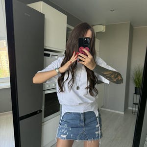 May include: A white t-shirt with a distressed denim skirt. The skirt has frayed edges and a light blue wash. The outfit is paired with white thigh-high stockings. The person is holding a black phone with a pink heart case.