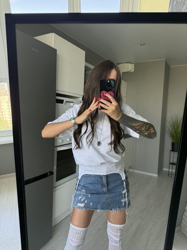 May include: A white t-shirt with a distressed denim skirt. The skirt has frayed edges and a light blue wash. The outfit is paired with white thigh-high stockings. The person is holding a black phone with a pink heart case.