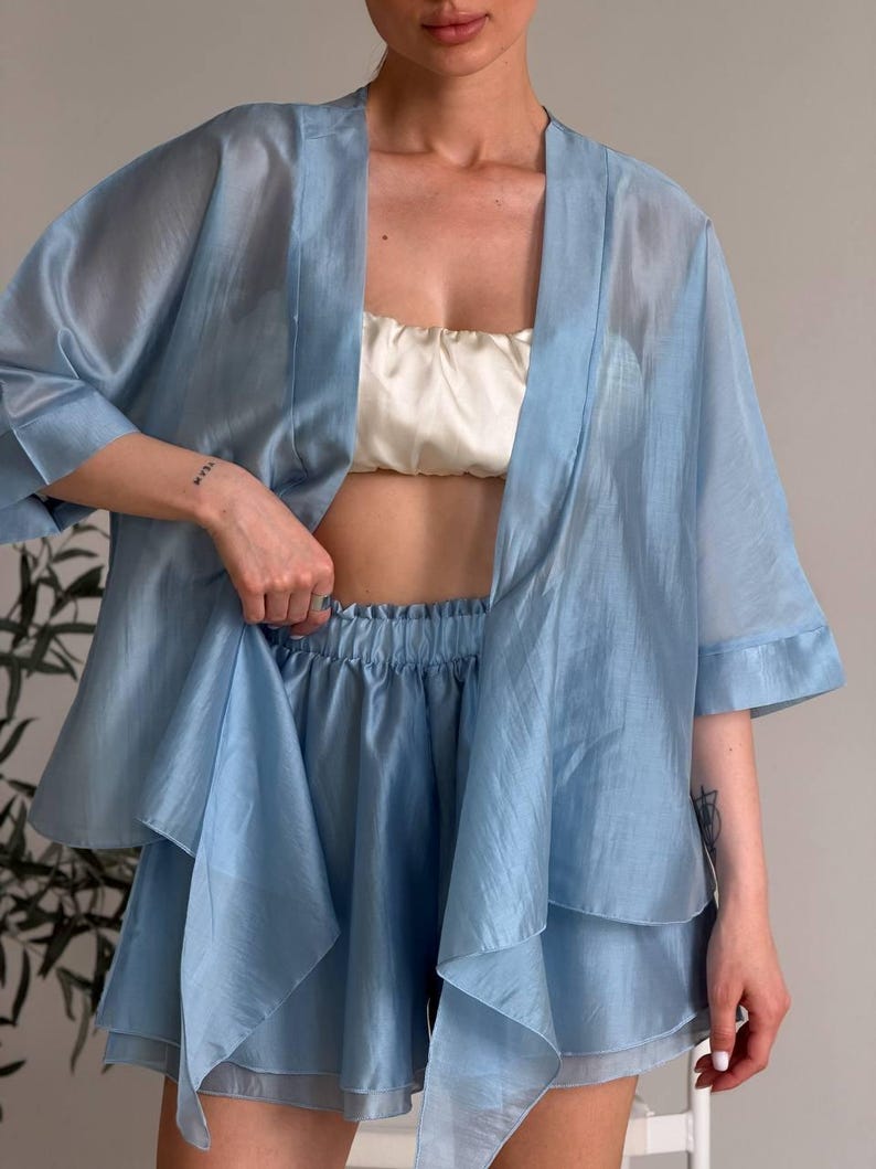 Op de afbeelding: Een lichtblauwe, doorschijnende, driedelige pyjamaset. De set bevat een kimono met korte mouwen en open voorkant, een bijpassende camisole en een short met ruches. De stof heeft een subtiele glans. De camisole is cr&egrave;mekleurig.