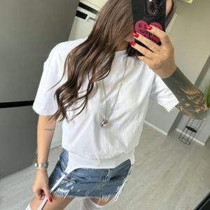 May include: White short-sleeved t-shirt paired with a denim mini-skirt. The skirt has distressed details and white suspender straps. The outfit is accessorized with a silver necklace and white thigh-high stockings.