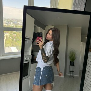 May include: A full-length mirror reflects a person wearing a white t-shirt, a distressed denim skirt, and white thigh-high socks. The mirror has a black frame. The background shows a kitchen and a cityscape.