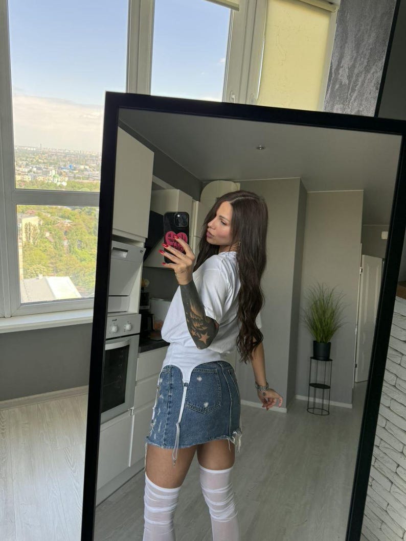 May include: A full-length mirror reflects a person wearing a white t-shirt, a distressed denim skirt, and white thigh-high socks. The mirror has a black frame. The background shows a kitchen and a cityscape.