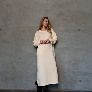 Puede incluir: Un vestido largo color crema con mangas largas y cuello redondo. El vestido tiene un corte holgado y un solo bolsillo lateral. La persona lleva botas negras. El fondo es una pared de hormigón gris.