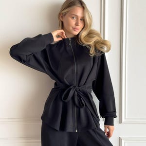 Puede incluir: Conjunto negro de dos piezas con una chaqueta con cremallera, cinturón anudado y pantalón de pernera ancha a juego. La chaqueta tiene mangas largas y cuello alto. El conjunto está hecho de una tela suave y mate, adecuado para ocasiones casuales o semiformales.