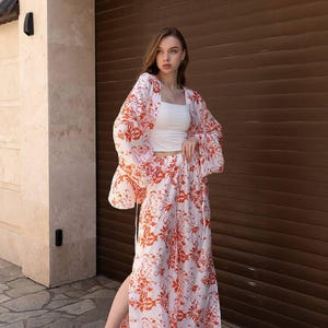 May include: A white two-piece outfit with a red floral pattern. The set includes a crop top, a long skirt with a side slit, and a kimono-style jacket with wide sleeves. The model is wearing white sandals.