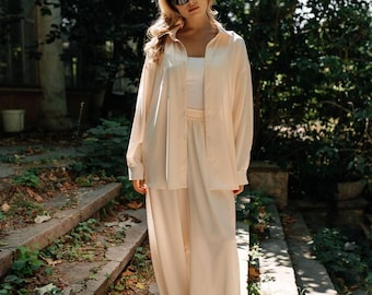 May include: A light peach-colored satin ensemble, including a button-down shirt, a tank top, and wide-leg pants. The outfit is worn with sunglasses. The model is standing on stone steps with greenery in the background. The outfit is suitable for casual or semi-formal occasions.