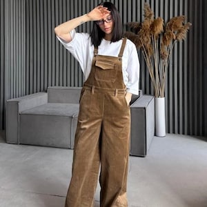 Puede incluir: Una persona con una camiseta blanca y un mono de pana marrón con un bolsillo delantero. El mono de pernera ancha tiene tirantes ajustables y se combina con zapatillas blancas. El fondo presenta un sofá gris y plantas decorativas.