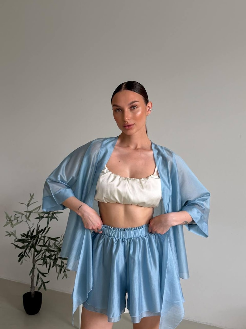 Op de afbeelding: Een lichtblauwe pyjamaset bestaande uit een kimono met korte mouwen, een camisole en een short. De camisole is wit. De pyjamaset is gemaakt van een lichte, vloeiende stof. Een kleine potplant staat op de achtergrond.