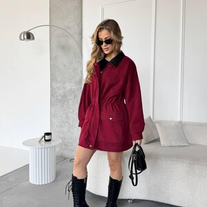 May include: A burgundy jacket dress with a black collar, cinched waist, and pockets. The outfit is paired with black combat boots and a small black handbag. The model is wearing sunglasses.