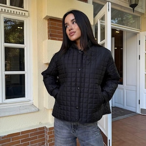 May include: A black quilted jacket with a button-up front and two pockets. The jacket has a diamond pattern and is worn with gray jeans. The person is standing outside near a building with a window and a door.