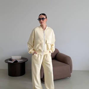 Puede incluir: Chándal amarillo claro que consta de una chaqueta bomber con cremallera y dos bolsillos delanteros y pantalones de pernera ancha. El conjunto se lleva con zapatillas blancas y gafas de sol. La chaqueta tiene cierre de cremallera.