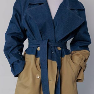 May include: A two-tone trench coat in denim blue and tan. The upper portion and sleeves are denim, while the lower part is tan. It features a matching belt, large lapels, and button closures. The coat has a classic trench coat design.
