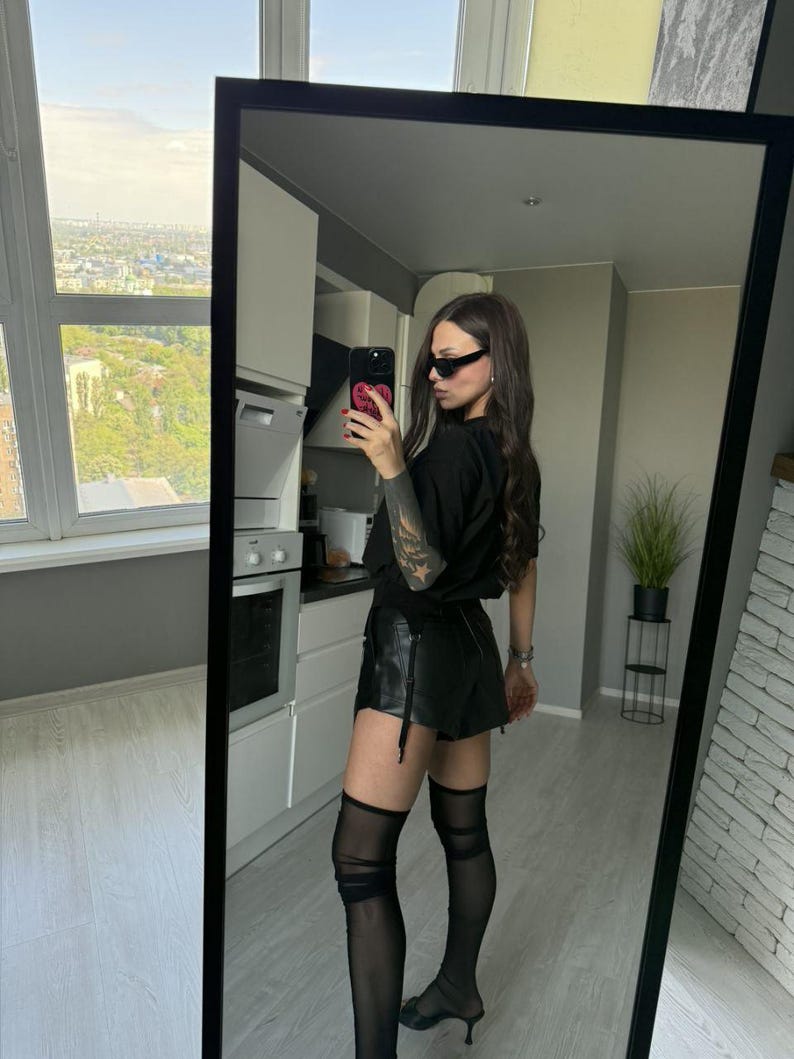 May include: A full-length mirror reflects a person wearing a black t-shirt, black leather shorts, and sheer black thigh-high stockings. The person is also wearing black sunglasses and black heels. The background includes a kitchen and a cityscape view.