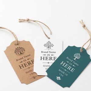 May include: Three rectangular paper tags with scalloped top edges and attached jute twine strings. One tag is brown, one is white, and one is dark teal. Each features a printed tree icon, the text Brand Name IN HERE, and MADE IN USA.