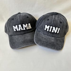 May include: Two black denim baseball caps with white embroidered text. One cap says "MAMA" and the other says "MINI".