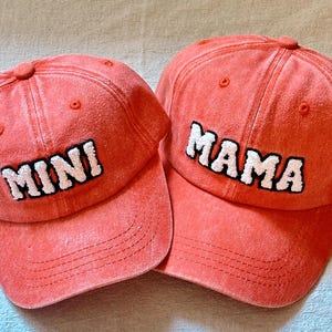May include: Two red baseball caps with white embroidered text. The larger cap says "MAMA" and the smaller cap says "MINI".