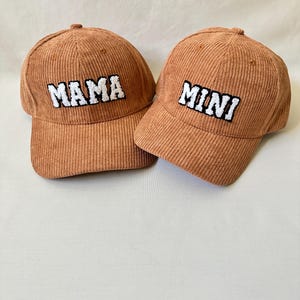 May include: Two corduroy baseball caps, one brown with "MAMA" embroidered in white, the other brown with "MINI" embroidered in white.