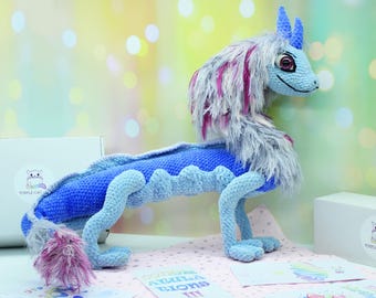May include: A blue and pink crocheted dragon plush toy with a long tail and a fluffy mane. The dragon has a friendly expression and is standing on a light pink surface.