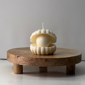 May include: A white candle shaped like a seashell with a pearl inside, sitting on a wooden pedestal.