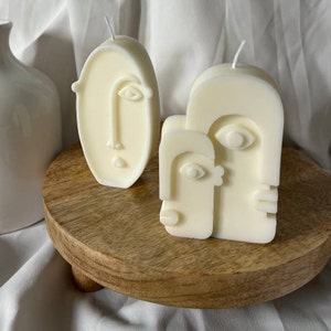May include: Three white candle sculptures in the shape of abstract faces. The candles are on a wooden pedestal.