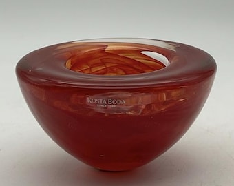 May include: A small, red glass candle holder with a swirl design. The holder is made by Kosta Boda, a Swedish glass company.