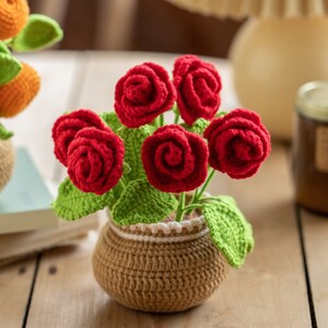 May include: A handcrafted crochet flower arrangement featuring six vibrant red roses with green leaves, nestled in a brown pot with a white trim. This decorative piece adds a touch of handmade charm to any space.