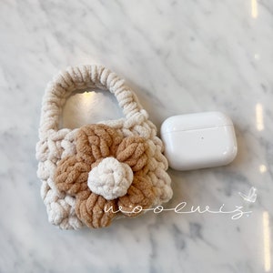 May include: A small, crocheted bag with a flower design in beige and brown. The bag has a handle and is sitting on a white marble surface. The bag is next to a white Airpods case.