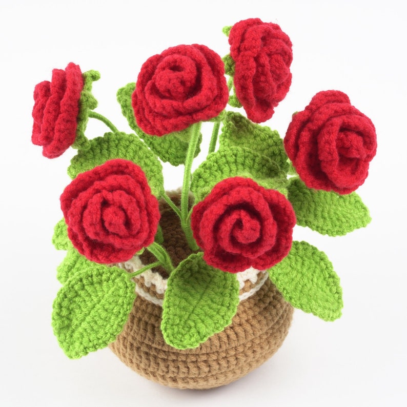 May include: A handcrafted crochet flower arrangement featuring vibrant red roses, green leaves, and a brown pot with a cream and brown trim. The roses are in full bloom, and the leaves are detailed. This decorative piece adds a touch of handmade charm.