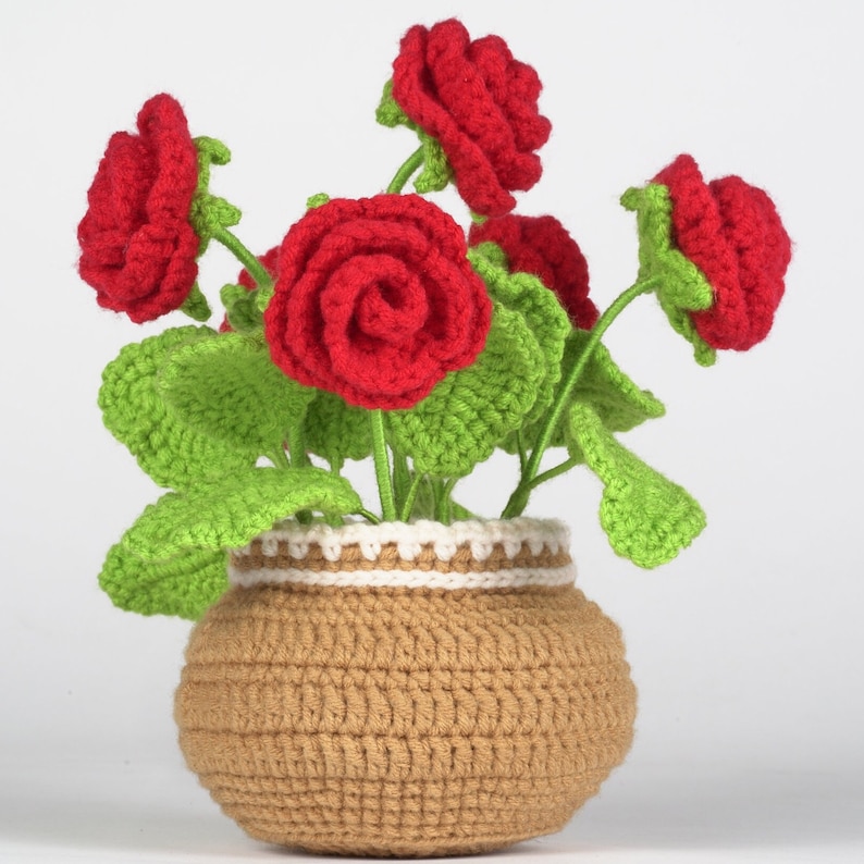 May include: A handmade crochet flower arrangement featuring vibrant red roses, green leaves, and a tan pot with white trim. The flowers are arranged in a decorative, handcrafted pot, creating a charming and colorful display. The crochet work is detailed and adds a unique touch.