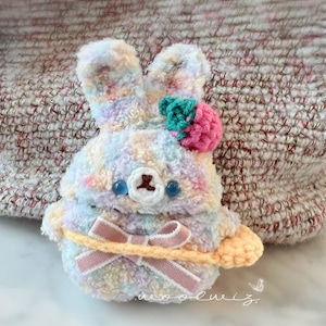 May include: A plush, pastel-colored bunny-shaped accessory with a bow and a flower accent. The bunny has a bear-like face with blue eyes and a light pink blush. The accessory is made of soft, fluffy material.