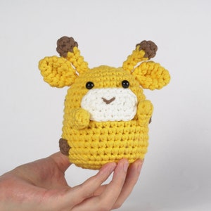May include: A crocheted stuffed giraffe toy with brown ears and black eyes. The giraffe is yellow and white and has a smiling face.