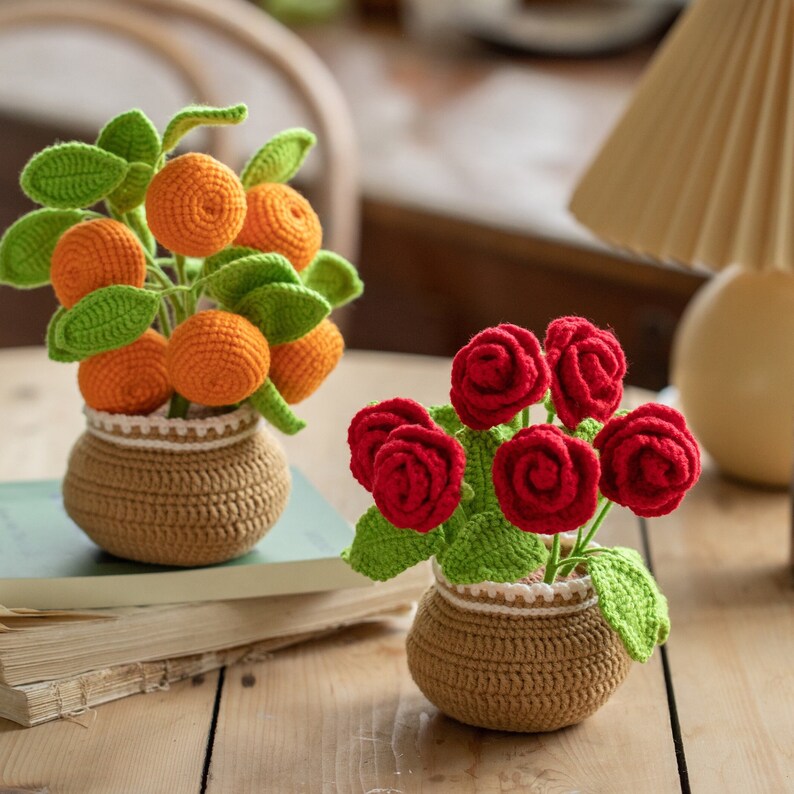 May include: Two crocheted flower arrangements. One features orange fruit with green leaves in a brown pot. The other has red roses with green leaves in a similar brown pot. Both pots have a white trim.