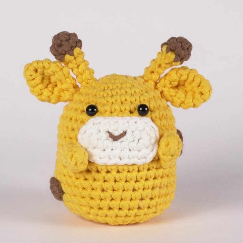 May include: A crocheted stuffed giraffe toy with brown ears, yellow fur, and black eyes. The giraffe has a white face with a stitched-on smile.