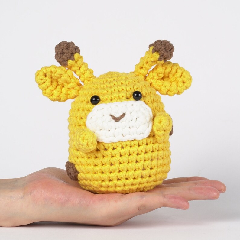 May include: A crocheted yellow giraffe toy with brown ears and black eyes. The toy is sitting in the palm of a hand.