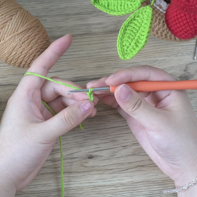 May include: Close-up of hands crocheting with green yarn and an orange-handled crochet hook. The project appears to be a leaf or other small crochet item. A ball of brown yarn and a partially completed red crochet item are in the background.