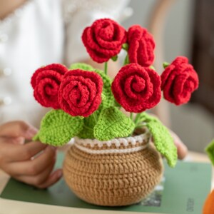 May include: A handcrafted crochet flower arrangement featuring vibrant red roses, green leaves, and a brown pot. The roses are in full bloom, and the pot has a decorative white trim. This charming piece is a unique home decor item.