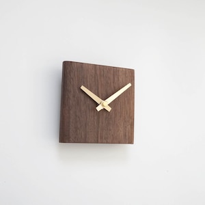 Puede incluir: Un reloj de pared cuadrado de madera con un acabado marrón oscuro. El reloj presenta manecillas doradas y un diseño minimalista. El reloj está montado en una pared blanca.