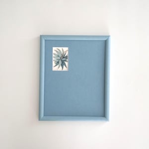 Könnte beinhalten: Ein gerahmter botanischer Druck mit einer blau-grünen Pflanze. Der Druck ist in einem hellblauen Rahmen auf weißem Hintergrund angebracht. Das Kunstwerk hat eine rechteckige Form und zeigt die Details der Pflanze.