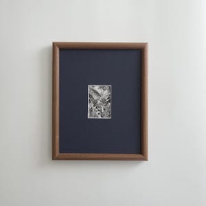 May include: A framed black and white artwork. The artwork is a small, detailed illustration set against a dark blue background. The frame is a warm brown color, and the artwork is mounted on a white wall.