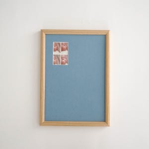 May include: A framed print with four small images of a baby's face. The frame is light-coloured wood, and the background is a solid, muted blue. The print is displayed against a white wall.