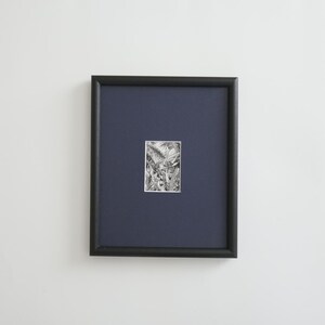 May include: A framed black and white artwork. The artwork is a detailed pen and ink drawing, set against a dark blue mat. The frame is a simple, black, rectangular shape. The artwork is centered within the frame, creating a clean and modern aesthetic.
