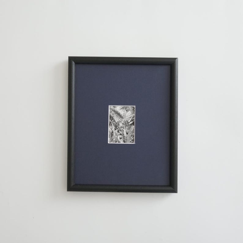 May include: A framed black and white artwork. The artwork is a detailed pen and ink drawing, set against a dark blue mat. The frame is a simple, black, rectangular shape. The artwork is centered within the frame, creating a clean and modern aesthetic.