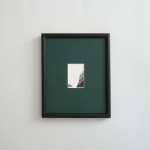 Op de afbeelding: Een ingelijst kunstwerk met een zwart frame, een donkergroene passe-partout en een kleine zwart-wit afbeelding. De afbeelding toont een deel van een gebouw. Het rechthoekige frame is aan een witte muur bevestigd.